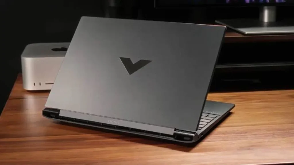 An HP Victus 15 laptop shown from the rear angle, displaying its matte dark grey lid with the reflective 'V' logo and a silver rear hinge bar on a wooden desk.