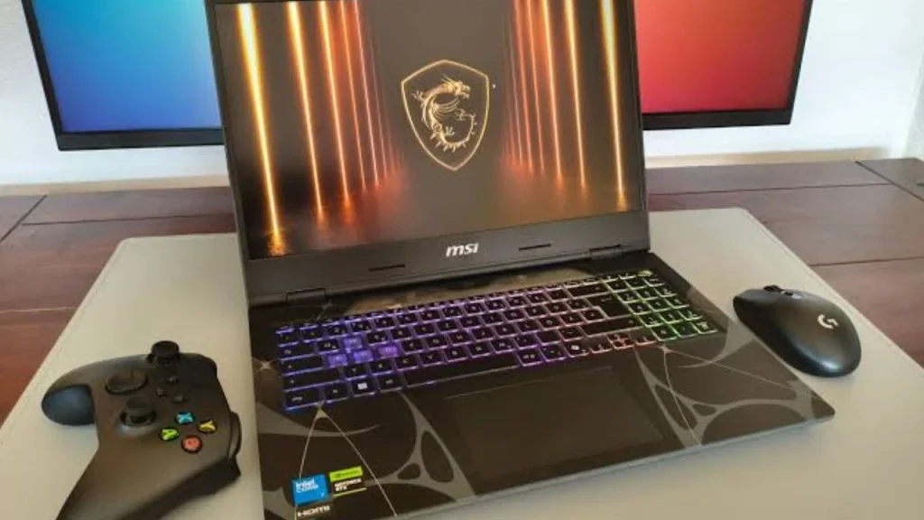 An open MSI Cyborg 15 gaming laptop on a light-colored desk mat, accompanied by a black gaming controller on the left and a black wireless gaming mouse on the right, with a dual-monitor backdrop.