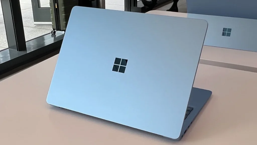 Top 10 Laptop Brands in 2026. A light blue Microsoft laptop viewed from the rear, showing the metallic lid with a centered, reflective Windows logo.