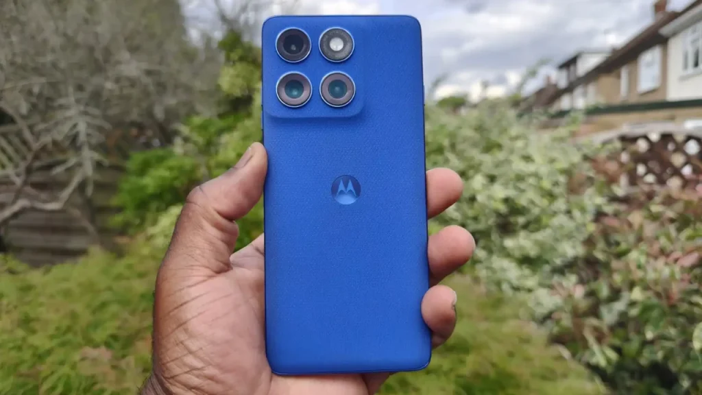 A hand holding a vibrant blue Motorola Edge 60 Pro smartphone with a textured back against an outdoor background of greenery.