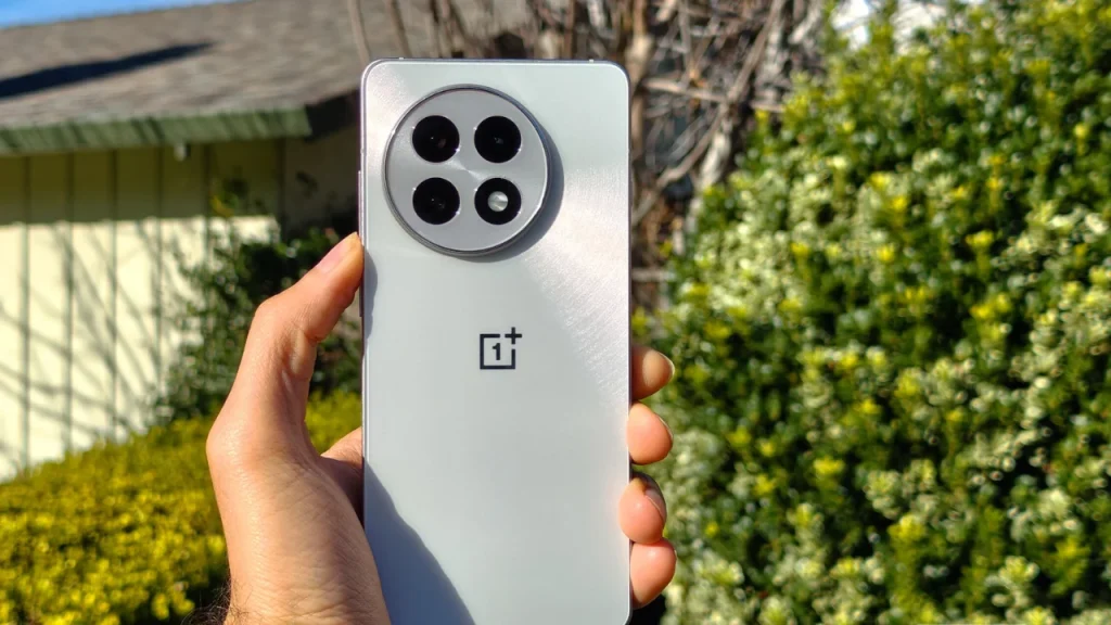 A person holding a silver OnePlus 13R smartphone outdoors with a blurred background of green foliage and a house roof.