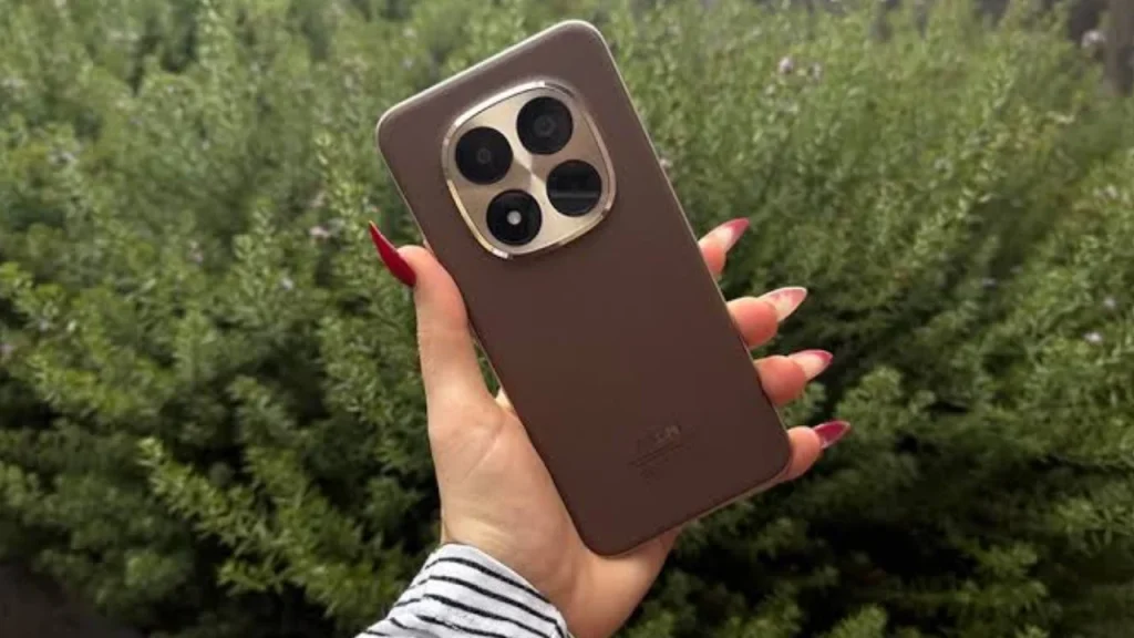 A hand with red-polished fingernails holding a brown Redmi Note 15 Pro Plus smartphone in front of a green bush.