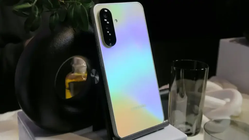 A Samsung Galaxy A36 5G with an iridescent, color-shifting back panel displayed upright on a stand next to a dark circular decorative object.