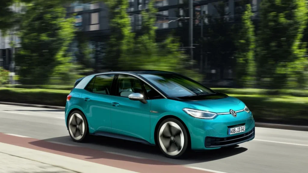 A vibrant teal Volkswagen electric hatchback driving down a city street lined with lush green trees, showcasing a practical choice among the Best EV brands.